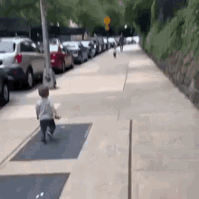 a baby is walking down a sidewalk next to a row of cars parked on the side of the road .