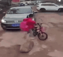 a man in a red jacket is pushing a child on a bike in front of a white car .