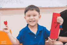 a young boy in a blue shirt is holding a piece of red paper with a heart cut out of it .