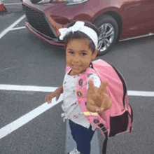 a little girl with a pink backpack is standing in a parking lot in front of a red car .