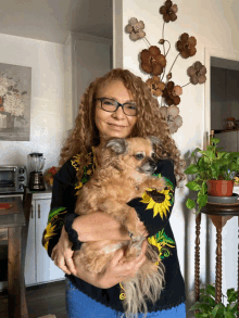 a woman in a sunflower sweater is holding a small brown dog