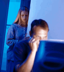 a woman stands behind a man who is using a computer