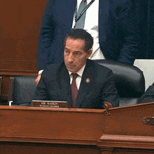 a man in a suit and tie is sitting at a desk with a name tag that says mr. raskin