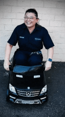 a woman in a blue shirt is sitting on a black mercedes benz toy car