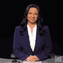 a woman in a blue suit is sitting at a desk with her hands folded in front of a snl sign .