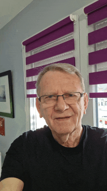 a man wearing glasses and a black shirt is smiling in front of a window with purple blinds