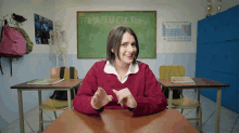 a woman in a red sweater sits at a desk in front of a blackboard that says toma tu cultura