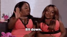 two women are sitting next to each other on a couch and one of them is saying `` sit down '' .