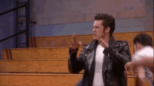 a man in a leather jacket is standing in front of a wooden bleachers and talking to a group of people .