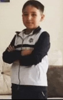 a young boy in a white and black jacket is standing with his arms crossed in a living room .