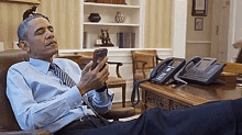 barack obama is sitting in a chair with his feet up looking at his phone