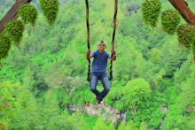 a man in a blue shirt is sitting on a swing in the middle of a forest