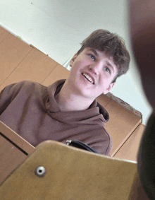 a young man in a brown sweatshirt sits at a desk smiling