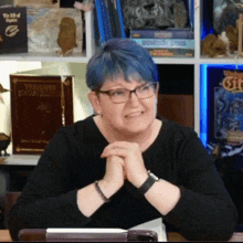 a woman with blue hair and glasses sits in front of a bookshelf with a book titled warhammer fantasy world