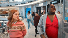 two women are standing next to each other in a shopping mall . one of the women is wearing a pink sweater .