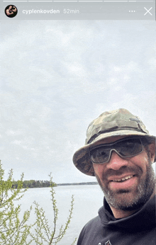 a man wearing a hat and sunglasses smiles in front of a body of water