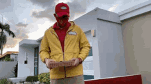 a man wearing a yellow jacket and a red hat is holding a pizza box in front of a house .