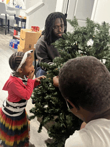 a man with dreadlocks is decorating a christmas tree while two children look on