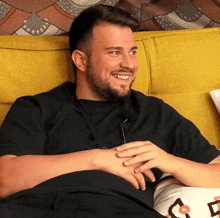 a man with a beard sits on a yellow couch smiling