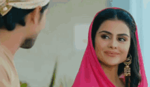 a man and a woman are looking at each other and smiling . the woman is wearing a pink scarf and earrings .