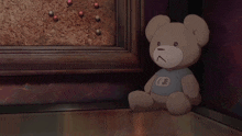 a teddy bear wearing a blue shirt with the letter k on it sits on the floor