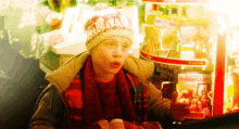 a young boy wearing a hat and scarf is sitting in front of a vending machine with a coca cola sign on it .