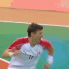 a man in a red and white shirt is running on a green field with the olympic rings in the background .