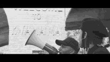 a black and white photo of a man holding a megaphone in front of a sign that says welcome to kamehameha