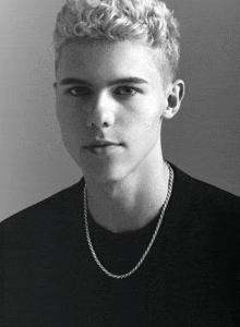 a black and white photo of a young man with curly hair wearing a chain around his neck