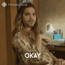 a man with long hair and a beard is sitting in front of a desk with the word okay on it