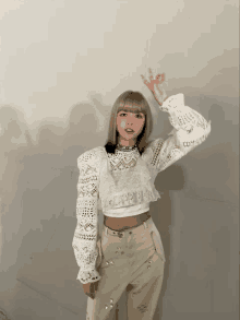 a woman wearing a white lace crop top and beige pants giving an ok sign