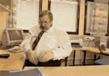 a man in a white shirt and tie is sitting at a desk in an office talking on a phone .