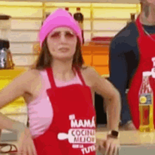 a woman in a pink hat and apron is standing in a kitchen with her hands on her hips .