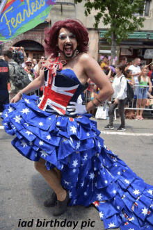 a drag queen in a red white and blue dress stands in front of a flag that reads fearless