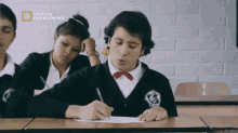 a boy in a school uniform is writing on a piece of paper in front of a national geographic sign