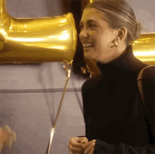 a woman wearing a black turtleneck is smiling in front of gold balloons hanging on a wall