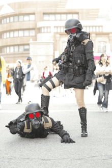 a woman in a gas mask stands next to a man in a gas mask laying on the ground