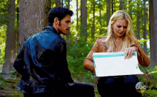 a man and a woman are sitting in the woods and the woman is holding a piece of paper with the abc logo on it