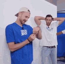 two men are standing next to each other in front of a white wall . one of the men is wearing a blue team edge shirt .