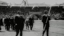 a black and white photo of a group of men walking in a stadium with the olympics logo in the background