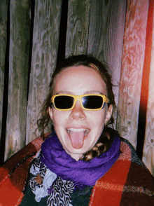 a woman wearing sunglasses and a plaid scarf sticks her tongue out