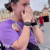 a woman in a purple shirt is covering her face with her hands while standing in front of a building .