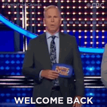 a man in a suit and tie is standing on a stage with the words welcome back written on the screen behind him .