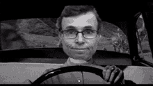 a man wearing glasses is driving a car and holding the steering wheel .