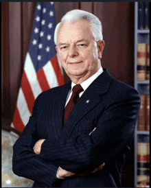 a man in a suit and tie with his arms crossed stands in front of an american flag