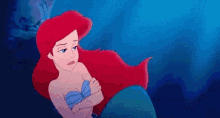 ariel from the little mermaid is sitting on the shore of the ocean with her arms crossed .