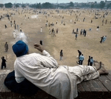 a man laying on a wooden ledge smoking a cigarette and looking at a crowd of people in a field