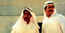 two men wearing white clothes and turbans are standing next to each other and looking at each other .