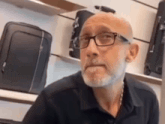 a bald man with glasses and a beard is sitting in front of a shelf full of luggage .