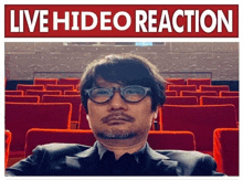 a man in a suit and glasses is sitting in an auditorium with the words live hideo reaction written above him